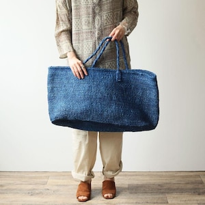 Könnte beinhalten: Eine große, rechteckige Tragetasche aus einem tiefblauen, strukturierten Webmaterial. Die Tasche verfügt über zwei passende blaue geflochtene Griffe, die an der Oberkante befestigt sind und einen festen Halt für dieses geräumige Accessoire bieten.