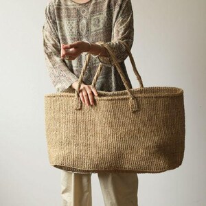 May include: A large, rectangular woven tote bag made from natural tan fibers. The bag features a wide, open top design with two sturdy, matching woven handles attached to the upper rim, providing a simple and functional structure for carrying items.