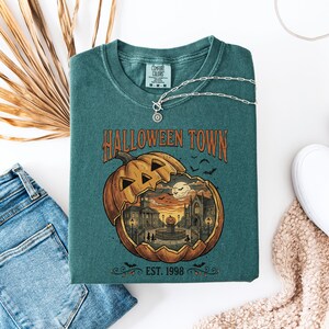 May include: A teal crewneck t-shirt featuring a graphic of a large carved pumpkin containing a town scene with buildings, a fountain, and a full moon. The text reads HALLOWEEN TOWN and EST. 1998. The shirt has a Comfort Colors label at the inner neckline.