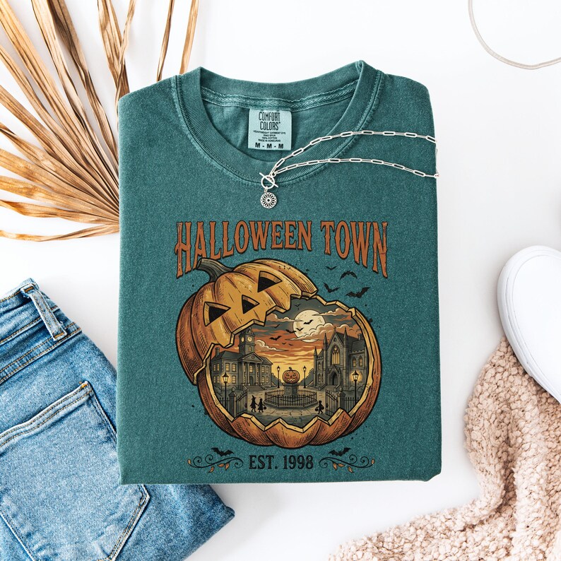 May include: A teal crewneck t-shirt featuring a graphic of a large carved pumpkin containing a town scene with buildings, a fountain, and a full moon. The text reads HALLOWEEN TOWN and EST. 1998. The shirt has a Comfort Colors label at the inner neckline.