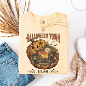 May include: A pale yellow t-shirt featuring a graphic of a large carved pumpkin revealing a town scene with buildings, a fountain, and bats. The text reads HALLOWEEN TOWN and EST. 1998. The shirt has a Comfort Colors brand label at the inner neckline.