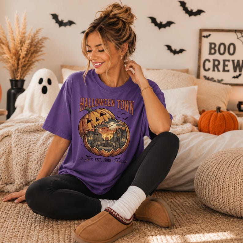 May include: A purple short-sleeved t-shirt featuring a graphic of a carved pumpkin revealing a spooky town scene inside. The text reads HALLOWEEN TOWN and EST. 1998. The design includes small bats and a full moon, printed on the front of the garment.