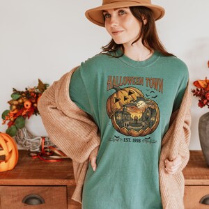 May include: A green t-shirt featuring a graphic of a carved pumpkin with a town scene inside, the text HALLOWEEN TOWN and EST. 1998, worn with a tan hat and a beige cardigan. The pumpkin design includes a full moon, buildings, and bats.
