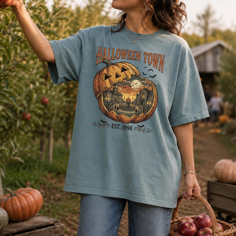 May include: A dusty blue short-sleeve t-shirt featuring a graphic of a carved pumpkin revealing a town scene inside. The text reads HALLOWEEN TOWN and EST. 1998, accompanied by small bat illustrations. The design is centred on the chest area.