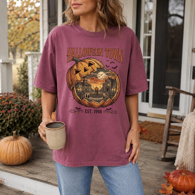 May include: A mauve short-sleeve t-shirt featuring a graphic of a large carved pumpkin with a town scene inside. The text reads HALLOWEEN TOWN and EST. 1998. The design includes small bats and a full moon, printed in orange, black, and cream tones.