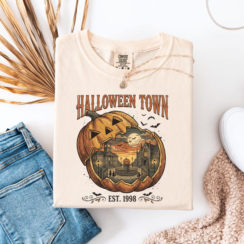 May include: A cream-coloured t-shirt featuring a graphic of a carved pumpkin with a town scene inside. The text reads HALLOWEEN TOWN and EST. 1998. The design includes a clock tower, buildings, a smaller pumpkin on a pedestal, and flying bats.