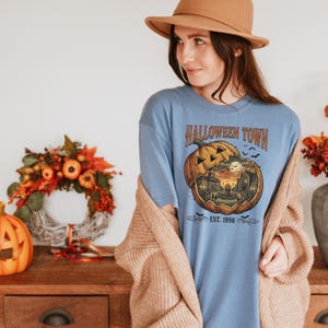May include: A blue short-sleeve t-shirt featuring a graphic of a carved pumpkin revealing a town scene inside. The text reads HALLOWEEN TOWN and EST. 1998. The design includes silhouettes of buildings, a fountain, and bats against a sunset sky.