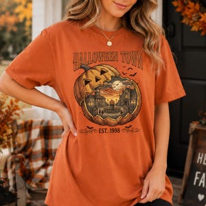 May include: An orange short-sleeve t-shirt featuring a graphic of a large carved pumpkin containing a town scene with buildings, a fountain, and the text HALLOWEEN TOWN EST. 1998. The design includes small flying bats and a full moon in the background.