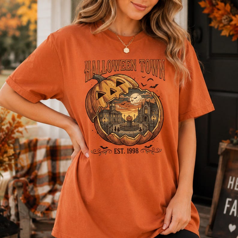 May include: An orange short-sleeve t-shirt featuring a graphic of a large carved pumpkin containing a town scene with buildings, a fountain, and the text HALLOWEEN TOWN EST. 1998. The design includes small flying bats and a full moon in the background.