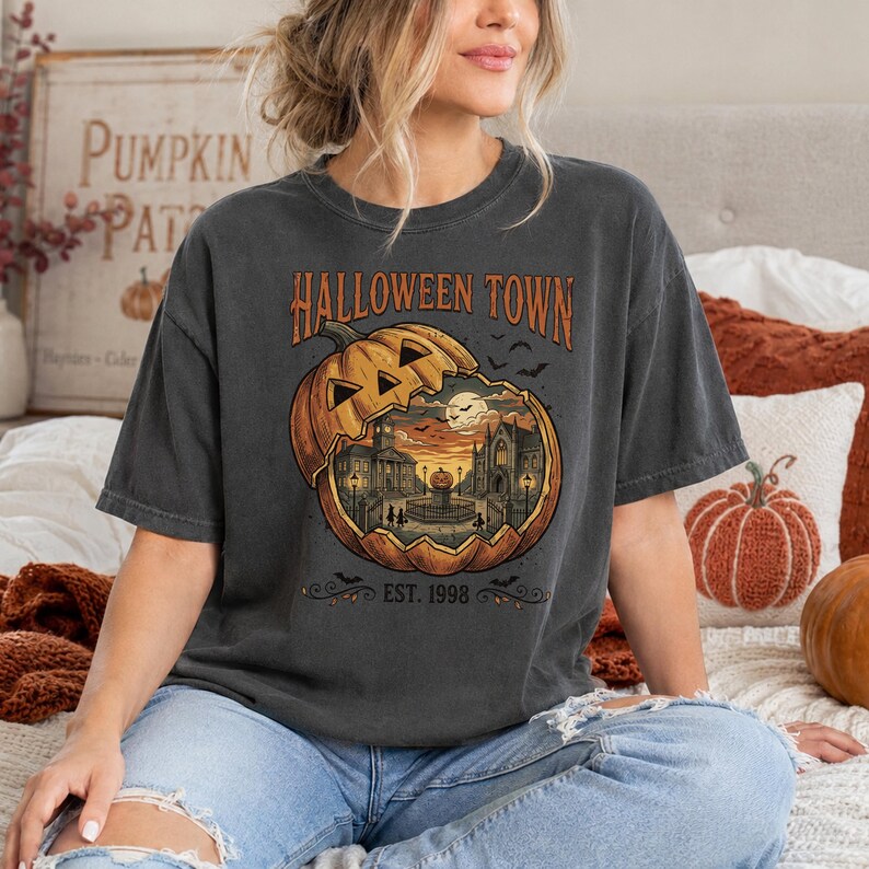 May include: A charcoal grey short-sleeve t-shirt featuring a graphic of a large carved pumpkin revealing a town scene inside. The text reads HALLOWEEN TOWN and EST. 1998. The design includes silhouettes of buildings, a full moon, and flying bats.