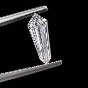 以下が含まれることがあります： 金属製のピンセットで保持された、シールド型の無色透明なファセットカットの宝石。石は尖った底部と平らな上部を持ち、内部で光が反射し、鋭く幾何学的なエッジが特徴で、高い透明度と輝きを放っています。