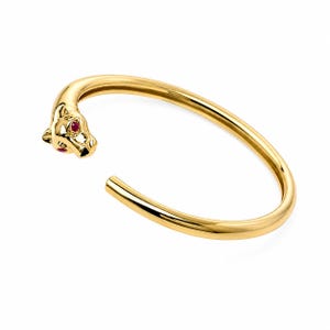 May include: A gold-toned open cuff bracelet featuring a sculpted panther head terminal at one end. The panther head is detailed with two small, vibrant red stones set as eyes, providing a distinct contrast against the polished metallic surface.