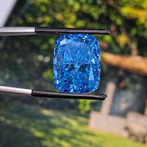 Peut inclure: Une pierre précieuse bleu vif taille coussin tenue par une pince en métal. La pierre présente une surface à facettes qui reflète la lumière, créant un aspect brillant avec des tons bleu clair et foncé dans toute la structure cristalline.