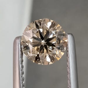 Könnte beinhalten: Ein runder, champagnerfarbener Edelstein mit Brillantschliff, der von zwei silberfarbenen Metallfassungen gehalten wird. Der Stein weist lichtreflektierende Oberflächen in warmen, bräunlich-beigen Tönen und eine symmetrische Form auf.