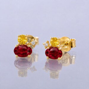 May include: A pair of 14k gold stud earrings featuring an oval-cut red gemstone, a round-cut yellow gemstone, and a small star-shaped accent with a clear stone. The gold posts are clearly marked with 14K, reflecting on a smooth surface.