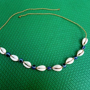 Puede incluir: Un collar compuesto por un cordón fino de color tostado, ensartado con conchas de cauri blancas y pequeñas cuentas redondas de color azul oscuro con diseño de ojo turco. Los elementos se disponen en un patrón alterno a lo largo del cordón.