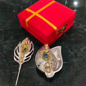 Puede incluir: Un bastón de metal plateado con forma de pluma de pavo real y un recipiente a juego con forma de hoja, ambos con detalles dorados, piedras transparentes y un motivo de ojo de pavo real. Detrás hay una caja de terciopelo rojo con una cinta amarilla.