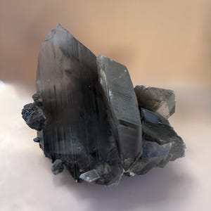 May include: A large, translucent smoky quartz crystal cluster featuring sharp, elongated points with vertical striations. The mineral specimen displays deep gray and charcoal tones, with smaller, jagged crystalline formations attached to the base.