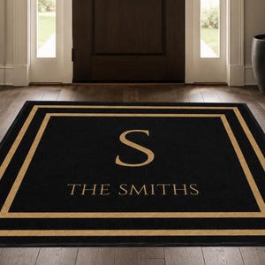 May include: A rectangular black floor mat featuring a gold-colored border design. The center of the mat displays a large gold letter S positioned above the text THE SMITHS in a matching gold font, creating a clear and centered visual arrangement.