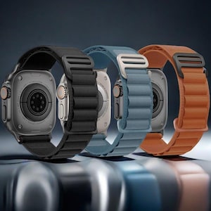 May include: Three smartwatches with metallic cases and textured nylon bands in black, blue, and orange. Each watch features a circular sensor array on the back, a side crown with an orange accent, and a rectangular metal buckle on the fabric strap.