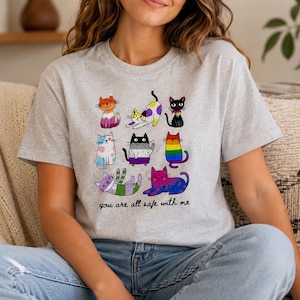 May include: A light grey crew neck t-shirt featuring a graphic of eight cartoon cats in various colours and patterns. Below the cats, the text "you are all safe with me" is printed in a simple black script font. The shirt has short sleeves.