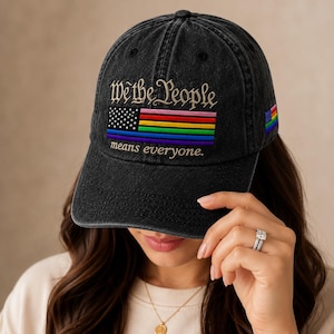May include: A black washed-denim baseball cap featuring embroidered text that reads We the People means everyone. Below the text is a rectangular graphic combining a black and white star field with horizontal rainbow stripes and a blue line.