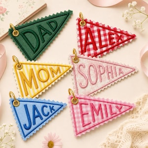 May include: Six triangular fabric pennants with scalloped edges and gold metal rings. The pennants feature embroidered names: DAD in green, ALEX in red gingham, MOM in yellow stripes, SOPHIA in pink floral, JACK in blue, and EMILY in pink gingham.