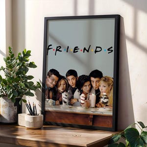 Op de afbeelding: Een ingelijste poster met het logo van de televisieserie Friends in het zwart met gekleurde stippen tussen de letters. Onder het logo zitten de zes hoofdrolspelers aan een bar, elk met een milkshake met een rietje in hun hand.