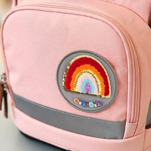 Könnte beinhalten: Ein hellrosa Rucksack mit braunen Kanten und dem Wort ergobag auf der oberen Klappe eingestickt. Die Vorderseite hat eine Reißverschlusstasche mit einem runden Aufnäher, der einen bunten Regenbogen und kleine Buchstabenperlen zeigt.