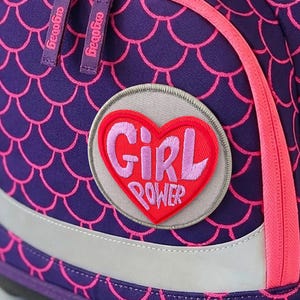 Può includere: Uno zaino viola con un motivo a squame di pesce rosa, una tasca frontale con doppia cerniera e una toppa circolare con un cuore rosso e la scritta Girl Power. La borsa presenta il logo ergobag azzurro e un dettaglio circolare giallo e rosa.