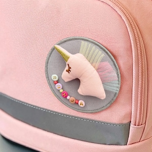 Può includere: Uno zaino rosa chiaro con il nome del marchio ergobag ricamato sulla patta superiore. La borsa include una chiusura con fibbia marrone, una tasca frontale con cerniera e una toppa circolare che mostra una testa di unicorno con il nome Ronja.