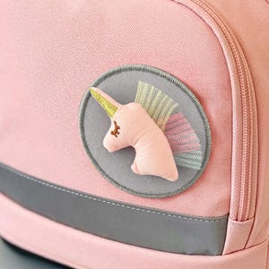 Peut inclure: Un sac à dos rose clair avec le nom de marque ergobag brodé en rose sur le rabat supérieur. Le sac comprend une fermeture à boucle marron, une poche avant zippée, une bande réfléchissante grise et un écusson circulaire avec un motif de licorne.