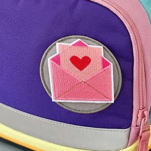 Könnte beinhalten: Ein mehrfarbiger Rucksack mit gelber Klappe und gesticktem ergobag-Schriftzug, einem türkisfarbenen Hauptteil und einer violetten Vordertasche. Eine orangefarbene Schnalle sichert die Klappe, ein runder Aufnäher zeigt einen rosa Brief mit Herz.