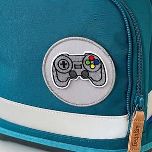 Può includere: Uno zaino color verde acqua con una patta superiore color cuoio che riporta la scritta ergobag. Lo zaino presenta un dettaglio circolare blu brillante, una tasca frontale con cerniera e una toppa rotonda con un controller di gioco grigio.