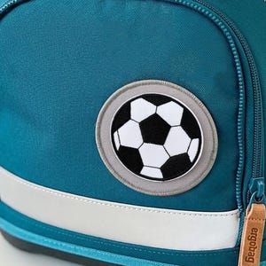 Peut inclure: Un sac à dos scolaire bleu sarcelle avec un rabat supérieur beige portant l'inscription ergobag en relief. Le sac comprend une fermeture circulaire bleu vif, une poche avant zippée avec un écusson de ballon de football et une bande réfléchissante.