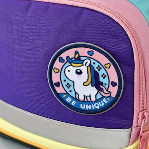Könnte beinhalten: Ein mehrfarbiger Rucksack mit hellvioletten, gelben und türkisfarbenen Einsätzen. Die Vordertasche ist dunkelviolett und mit einem runden Aufnäher versehen, der ein weißes Einhorn und den Text BE UNIQUE zeigt. Eine orangefarbene Schnalle mit ergobag-Logo.