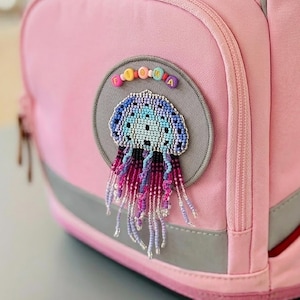 Può includere: Uno zaino rosa e rosso con la scritta ergobag stampata sulla patta superiore. La tasca frontale presenta una toppa circolare grigia decorata con un design a medusa in perline nei toni del blu e del viola, con perline a lettere colorate LOUKA.