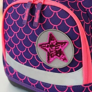 Könnte beinhalten: Ein violetter Schulrucksack mit pinkem Fischschuppenmuster, einer Vordertasche mit pinken Reißverschlüssen und einem runden Aufnäher mit einem pinken Paillettenstern. Der Markenname ergobag ist hellblau auf den Deckel gestickt, ergänzt durch einen gelben Ring.