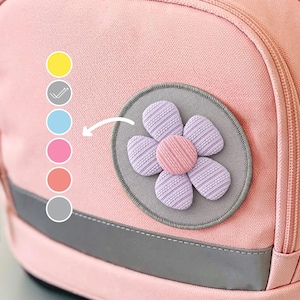 Könnte beinhalten: Ein hellrosa Rucksack mit dem Schriftzug ergobag auf der oberen Klappe und einem Schnallenverschluss. Auf der Vordertasche befindet sich ein kreisförmiger Aufnäher mit einer lavendelfarbenen Blume sowie eine vertikale Reihe aus sechs farbigen Kreisen.