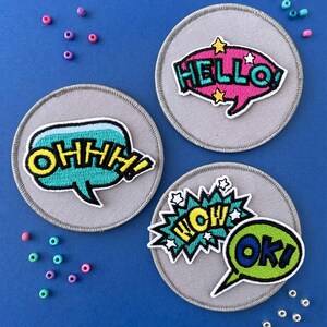 Könnte beinhalten: Drei runde graue Stoffaufnäher mit gestickten Sprechblasen. Ein Aufnäher zeigt eine pinke Sprechblase mit dem Text HELLO, ein weiterer eine türkise Sprechblase mit OHHH und der dritte eine türkise WOW-Explosion sowie eine limettengrüne OK-Sprechblase.