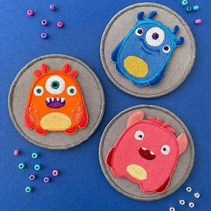 May include: Three circular embroidered patches featuring cartoon monsters on a gray background. One patch shows an orange monster with three eyes, one shows a blue monster with one eye, and one shows a pink monster with two eyes and a yellow belly.