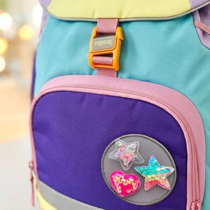 Könnte beinhalten: Ein mehrfarbiger Rucksack mit gelber Klappe und dem Schriftzug ergobag, einem türkisfarbenen Hauptteil und einer violetten Vordertasche. Die Vordertasche zeigt einen runden Aufnäher mit einem transparenten Stern voller Konfetti, einem rosa Paillettenherz und einem türkisfarbenen Paillettenstern.