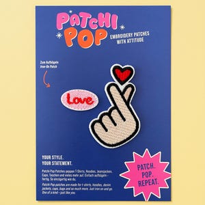 Könnte beinhalten: Zwei gestickte Aufbügel-Patches auf einer blauen Trägerkarte. Ein Patch ist oval mit dem Wort Love in roter Schrift. Der andere Patch zeigt eine Handgeste, die ein Herz formt, mit einem kleinen roten Herz darüber. Der Text umfasst Patchi Pop, Embroidery Patches With Attitude und Patch. Pop. Repeat.