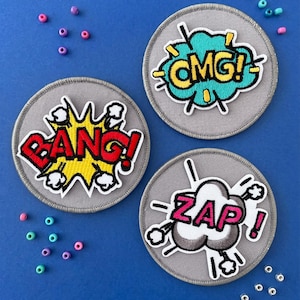 Puede incluir: Tres parches bordados circulares de color gris con efectos de sonido de cómic. Un parche muestra la palabra BANG en rojo, otro OMG en amarillo y el tercero ZAP en rosa. Cada parche está rodeado de cuentas de colores dispersas.