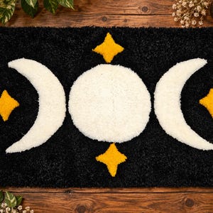 May include: A rectangular tufted rug with a black background featuring a large white central full moon flanked by two white crescent moons. Four yellow four-pointed stars are positioned above, below, and to the sides of the central moon.