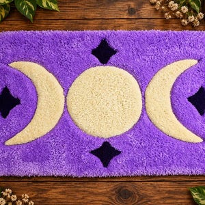 May include: A rectangular purple tufted rug featuring a central cream-colored full moon flanked by two cream-colored crescent moons. The design includes four dark purple four-pointed stars positioned above, below, and to the sides of the moons.