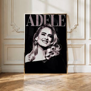 May include: A black and white portrait poster of the singer Adele, featuring the name ADELE in large capital letters at the top. The image shows a smiling person with wavy hair and a dark earring, set against a dark background within a gold frame.