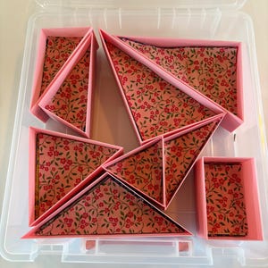 Puede incluir: Un contenedor de almacenamiento de plástico transparente que contiene varias cajas de paredes rosas de diversas formas geométricas, incluyendo triángulos y rectángulos. Cada caja está forrada con una tela rosa con un patrón de pequeñas flores rojas y hojas verdes.