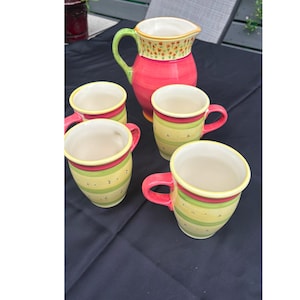 May include: A ceramic pitcher and four matching mugs featuring a vibrant color palette of pink, lime green, and yellow. The pitcher has a floral pattern around the rim, while the mugs display horizontal stripes and small dot accents throughout.
