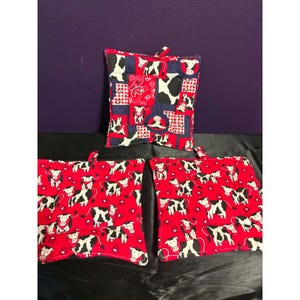 May include: Three square fabric pot holders featuring a red, white, and black cow print pattern. Two pot holders display a repeating cow motif on a red background, while the third features a patchwork design of cows, paisley, and checkered squares.